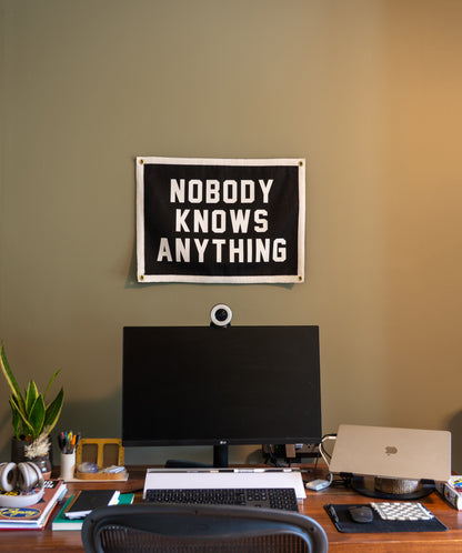 Home office setup with a computer monitor, keyboard, and 'Nobody Knows Anything' flag on the wall.
