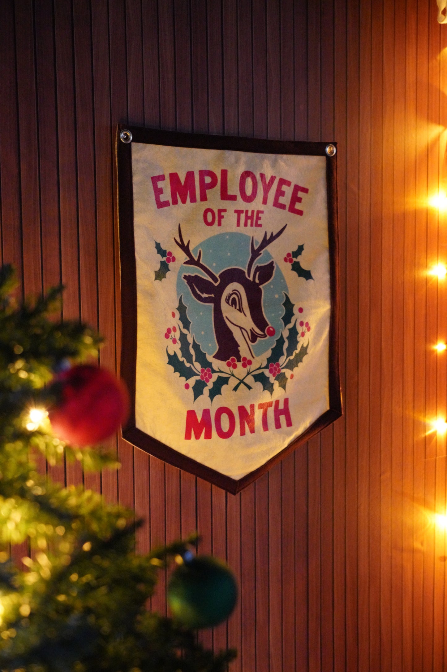 Decorative banner with 'Employee of the Month' design hanging on a wooden wall with Christmas tree and lights.