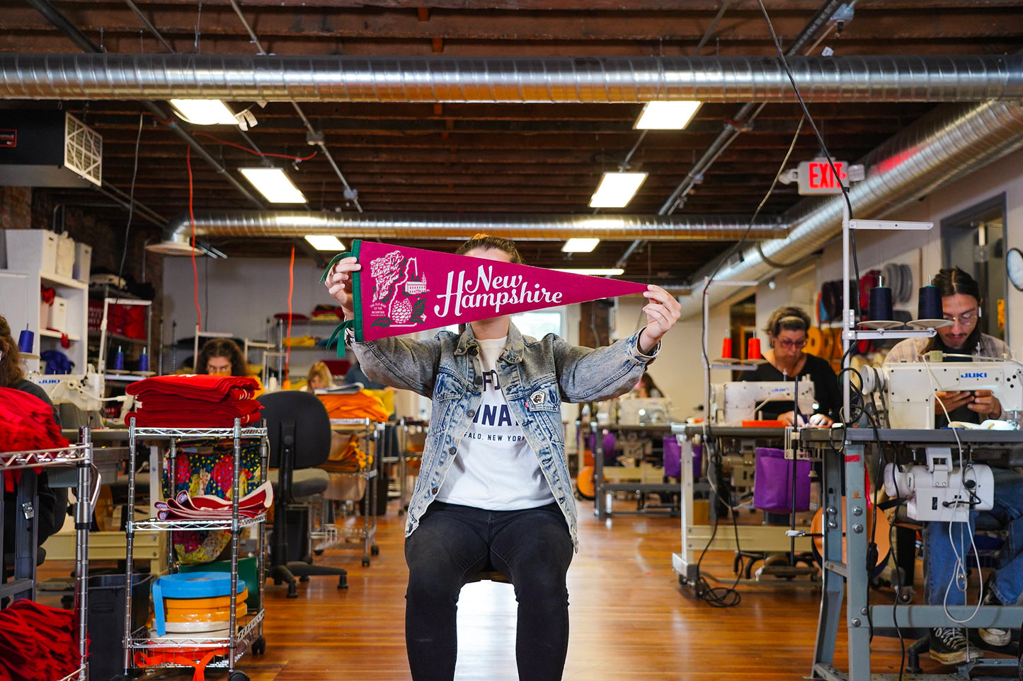 Person holding a New Hampshire sign in a busy workshop with sewing machines and materials.